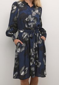 Navyblauwe jurk met abstract grijs en zwart bloemenpatroon, knoopsluiting aan de voorkant, lange pofmouwen, strikceintuur op de taille en zijzakken.