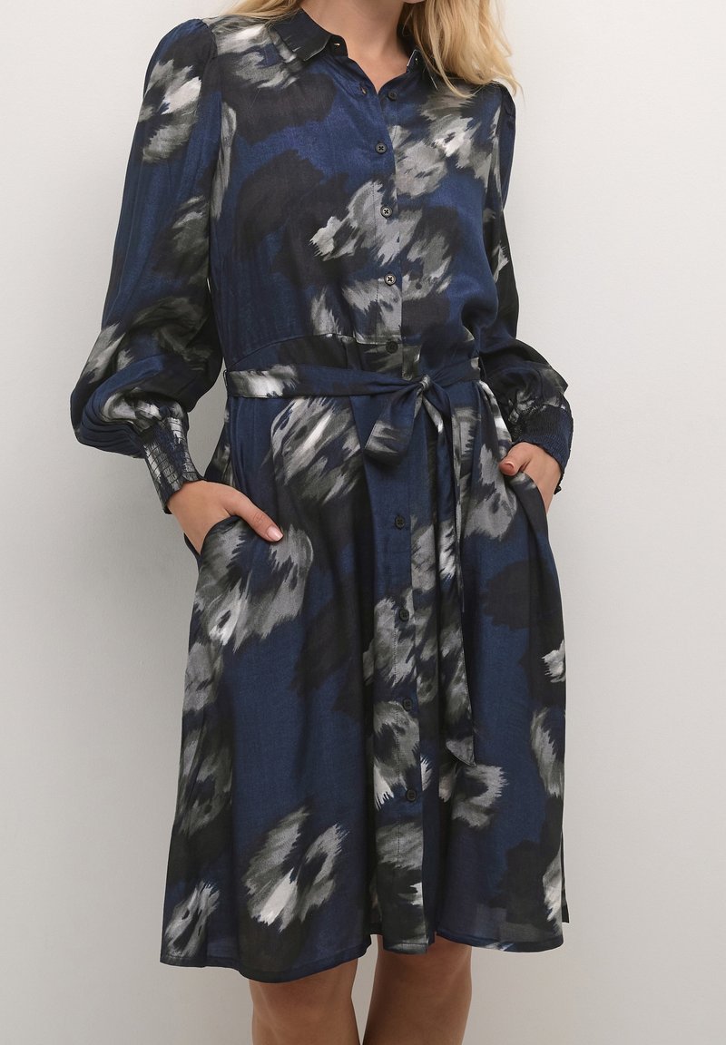 Navyblauwe jurk met abstract grijs en zwart bloemenpatroon, knoopsluiting aan de voorkant, lange pofmouwen, strikceintuur op de taille en zijzakken.