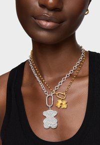 Des colliers superposés en argent et en or comprenant un pendentif ours en argent, des charms fleurs en or, et des chaînes à maillons entrelacés de tailles variées.