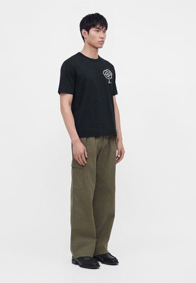 Homme debout portant un t-shirt noir avec un petit motif floral blanc et un pantalon cargo ample vert olive avec des chaussures noires.
