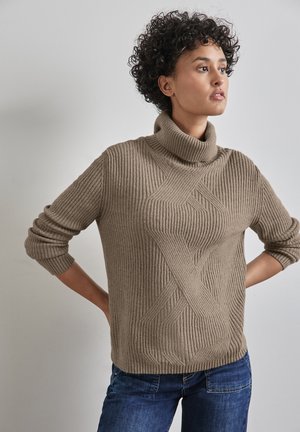Beżowy sweter typu golf z prążkowanym wzorem i skomplikowaną teksturą. Ma luźny krój i jest zestawiony z niebieskimi dżinsami.