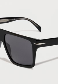 Melnas taisnstūra saulesbrilles ar biezām rāmjiem un tumšiem stikliem, attēlotas tuvplānā uz baltas fona.