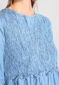 Sininen farkkumekko, jossa on teksturoitu, smockattu yläosa, pyöreä kaula-aukko ja revitty helma. Valmistettu kevyestä farkkukankaasta.