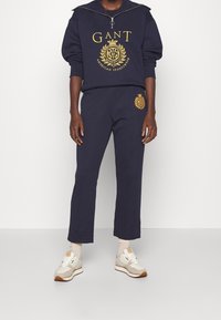 Sudadera azul marino con mangas oversized y detalle del logo en dorado, combinada con pantalones de chándal azul marino a juego que presentan una marca bordada. Incluye zapatillas.
