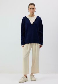 Tmavomodrý nadrozmerný sveter s krémovým výstrihom do V a dvoma gombíkmi, v kombinácii s voľnými béžovými nohavicami a bielymi teniskami. Jednoduchý dizajn.