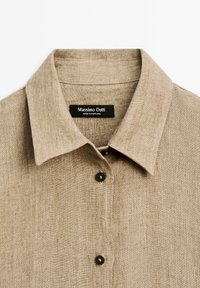 Beige linnedkjorte med krave, to mørke knapper og en synlig sort mærkat med teksten "Massimo Dutti Made in Portugal."
