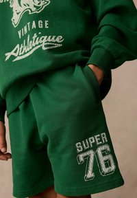Grön sweatshirt och shortset i bomull, med vita tryck: "VINTAGE ATHLETIQUE" och "SUPER 76" med texturerad detaljering.