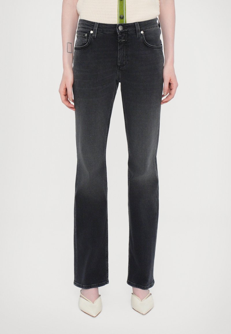 Femme portant un jean droit noir avec une fermeture à bouton et zip, associé à des talons blancs à bouts pointus.