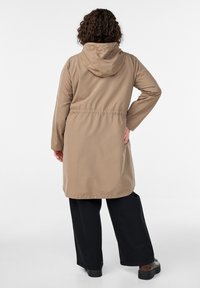 Beiger, wasserdichter Mantel mit Kapuze, elastischer Taille, langen Ärmeln und lockerem Sitz. Kombiniert mit weiten schwarzen Hosen und klobigen schwarzen Schuhen.