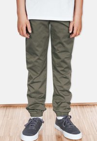 Olivgröna tapered byxor med elastiska muddar, tillverkade av mjukt tyg. Matchade med mörka sneakers med vita sulor och snören.