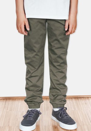 Olivgrüne tapered Hosen mit elastischen Bündchen, aus weichem Stoff. Kombiniert mit dunklen Sneakern mit weißen Sohlen und Schnürsenkeln.