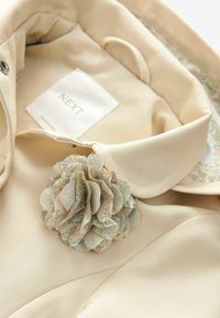 Col de veste couleur crème avec une broche fleur en tissu floral et une étiquette de marque blanche "NEXT" à l'intérieur.
