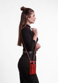 Červená prošívaná kožená kabelka přes rameno s řetízkovým popruhem, která má přední kapsu a zlaté kovové detaily, předvedená na modelu v profilu.