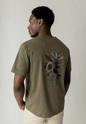 Uomo visto di profilo che indossa una maglietta verde oliva con un motivo floreale e la scritta "Mind in Motion" sul retro, abbinata a pantaloni di colore chiaro.