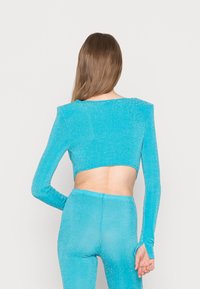 Top cropped turquesa com mangas longas e recorte nas costas, feito de um tecido brilhante. Apresenta um design justo e uma textura suave.