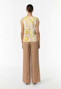 Kurzärmliges Oberteil aus leichtem Stoff mit einem gelb-beigen abstrakten Muster; kombiniert mit lockeren, hochgeschnittenen beigen Hosen. Elastische Ärmelbänder.