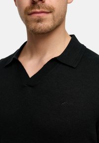 Schwarzes, gestricktes Polohemd mit V-Ausschnitt und dezentem Logo auf der Brust. Der Stoff erscheint glatt und leicht strukturiert.