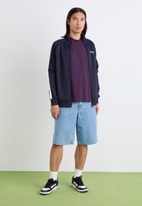 Marine glidelåsjakke, lilla skjorte, lyseblå denimshorts, svart og hvit sneakers, står på grønt gulv mot en enkel bakgrunn.