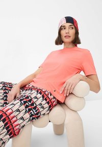 Coral short-sleeve t-shirt with a round neckline paired with patterned pants in pink, red, and black geometric shapes. Model poses seated.