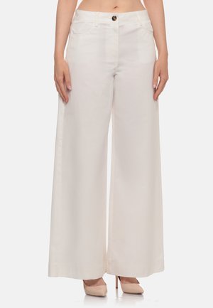 Donna che indossa pantaloni bianchi a vita alta e gamba ampia con bottone frontale e tasche, abbinati a scarpe beige con tacco alto, in piedi.