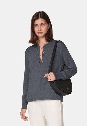 Graues Langarmhemd mit Henley-Ausschnitt und dekorativen Knöpfen, kombiniert mit einer schwarzen Tasche mit gekrümmtem Schulterriemen und hellen Hosen.