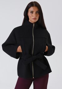 Giacca nera con zip e colletto alto, maniche ampie e cintura legata. Presenta una zip dorata e tasche frontali. Tessuto morbido e testurizzato.