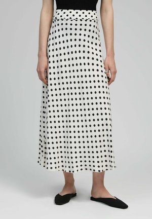 Jupe maxi blanche à pois noirs, texture lisse, taille haute, avec un design simple et une coupe droite. Accessoirisée avec des chaussures noires à enfiler.