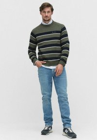 Groen en marineblauw gestreepte trui met een ronde hals, gedragen over een lichtblauw shirt, gecombineerd met lichtblauwe jeans en donkere sneakers.