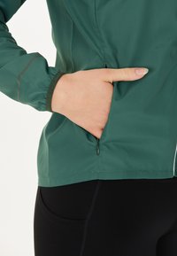 Groene windjack met een ritszak opzij, elastische manchetten en een gladde textuur. Draagt zich met zwarte leggings met zijzakken.