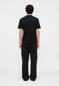 Άνδρας με σγουρά μαλλιά στέκεται γυρισμένος με την πλάτη φοράει απλό μαύρο μπλουζάκι και φαρδιά μαύρη παντελόνια μπροστά σε λευκό φόντο.