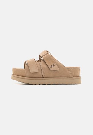 Sandalias deslizantes de ante beige con dos correas ajustables y una suela texturizada que presenta un ligero tacón y un detalle de logo distintivo.