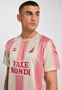 Koszulka z krótkim rękawem w jasnobeżowym kolorze z różowymi pionowymi paskami. Z kolorowym logo i napisem "FAKE KONDI" w odważnym różowym kolorze. Materiał o fakturze.