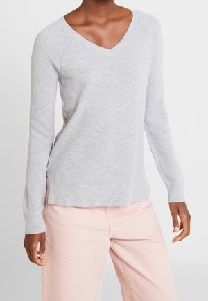 Personne portant un pull à col en V texturé gris clair et un pantalon rose pâle, debout devant un fond blanc uni.