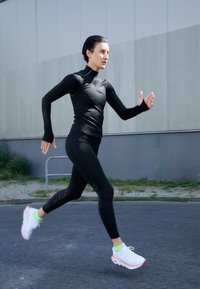 Czarna, długorękawowa koszulka sportowa z otworami na kciuki, w połączeniu z dopasowanymi czarnymi legginsami. Ubranie ma teksturowane akcenty. Białe sneakersy z neonowo zielonymi skarpetami.