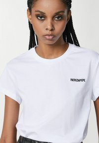 T-shirt bianca in cotone con maniche corte, collo rotondo, caratterizzata da un logo ricamato nero "PATRIZIA PEPE" sulla parte sinistra del petto.