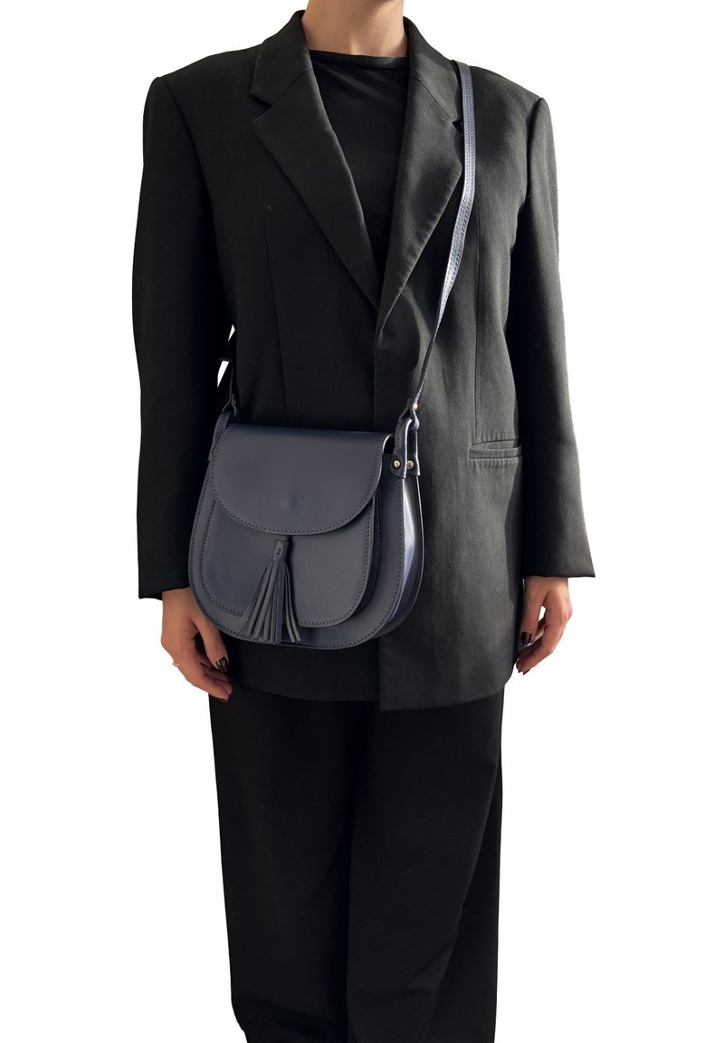 Kobieta w czarnym żakiecie i spodniach, nosząca ciemnoniebieską skórzaną torebkę crossbody z frędzlem na przedniej klapie.