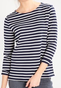 Žena v námořnickém modrém a bílém pruhovaném dlouhém tričku s lodní výstřihem a šedých kalhotách, stojící na jednobarevném pozadí.