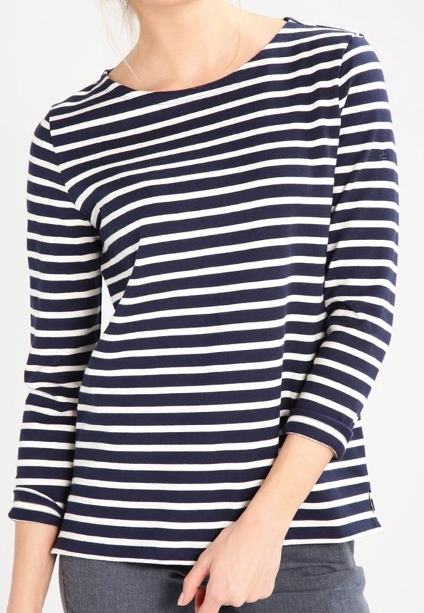 Žena v námořnickém modrém a bílém pruhovaném dlouhém tričku s lodní výstřihem a šedých kalhotách, stojící na jednobarevném pozadí.