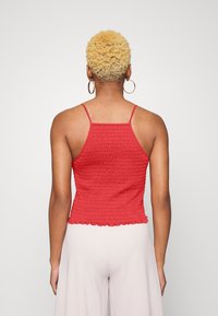 Top de tanque vermelho pregueado com alças finas, apresentando uma superfície texturizada e uma bainha com babados. Exibido sobre um fundo neutro.
