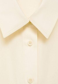 Camicia button-up beige chiaro con un colletto grande e strutturato e quattro bottoni rotondi. Realizzata in un tessuto morbido.