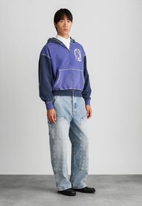 Mikina s kapucňou v modrom prechode s predným zipsom a bielou grafiku astronauta; spojená s voľnými, svetlými denimovými džínsami.