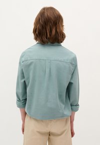 Chemise à manches longues bleu clair, avec un col et des manches retroussées, dotée d'une couture centrale dans le dos, fabriquée à partir d'un tissu lisse.