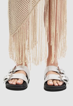 Nohy v stříbrných sandálech na přezku pod béžovým třásňovým pleteným oděvem na jednolitém pozadí.