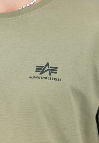 Olivgrünes T-Shirt aus weichem Baumwollstoff, mit einem schwarzen Logo von "ALPHA INDUSTRIES", das ein stilisiertes "A" und horizontale Streifen zeigt.