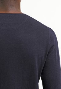 Homme vu de dos portant un pull à manches longues bleu marine foncé avec des coutures visibles sur l'épaule et le col.