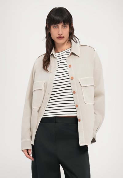 Jachetă bej deschis cu două buzunare frontale, închidere cu nasturi și croială lejeră, purtată peste un top în dungi alb-negru.