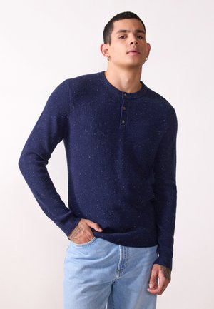 Navy knitted long-sleeve shirt with a crew neckline, three buttons at the collar, and speckled texture; paired with light blue jeans.