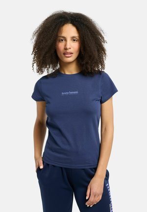 Kvinde med krøllet hår iført en tætsiddende mørkeblå t-shirt og mørkeblå bukser, stående med den ene hånd i lommen mod en hvid baggrund.