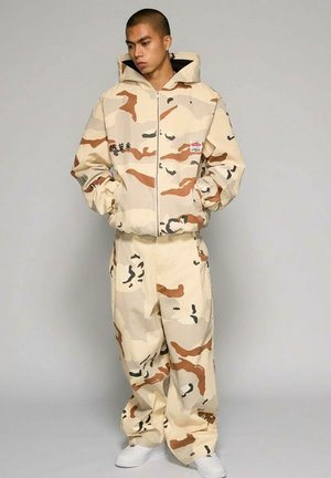 Man draagt een beige, bruin en zwart camouflage gewatteerde jas met capuchon en bijpassende losse broek, staand met handen in de zakken, witte sneakers.