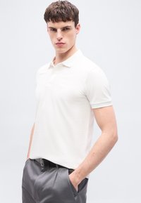 Νέος άνδρας με κοντά σγουρά μαλλιά, φοράει λευκό πόλο μπλουζάκι και γκρι παντελόνι, με τα χέρια στις τσέπες, μπροστά σε απλό φόντο.